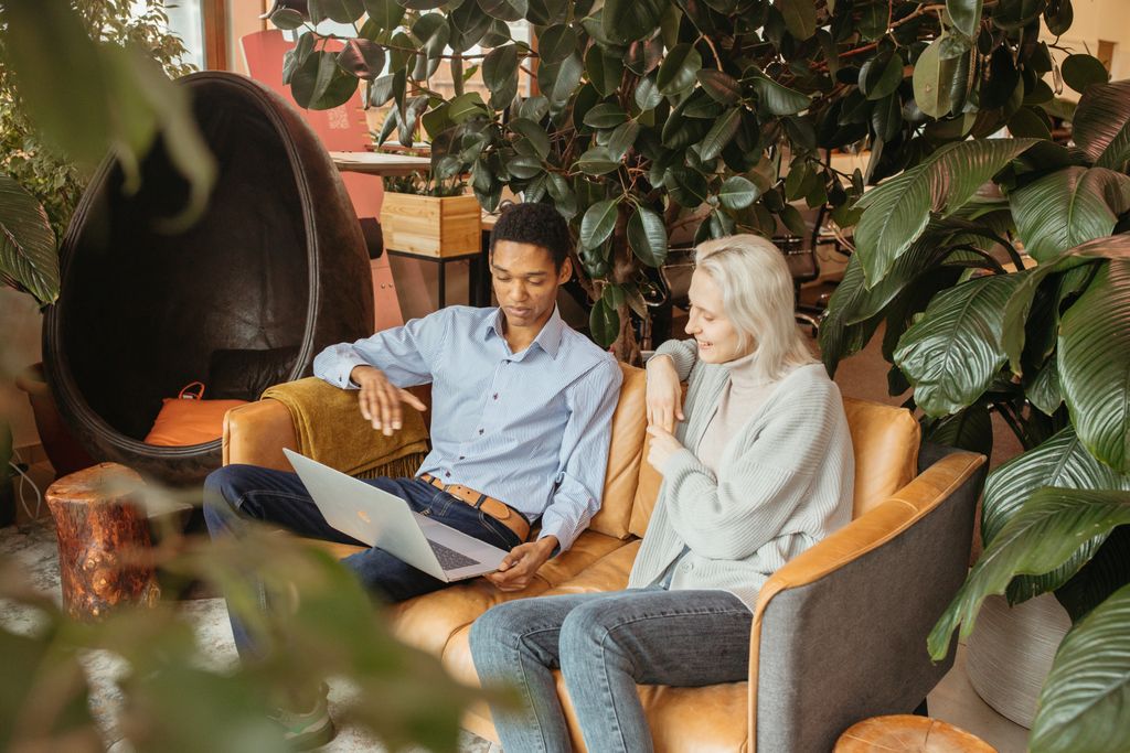 Two coworkers sitting on a sofa, working together using a laptop surrounded by indoor plants.